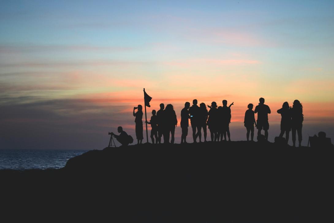 Silhouette of a group of people at sunset near a coastline. One person appears to be holding a flag.